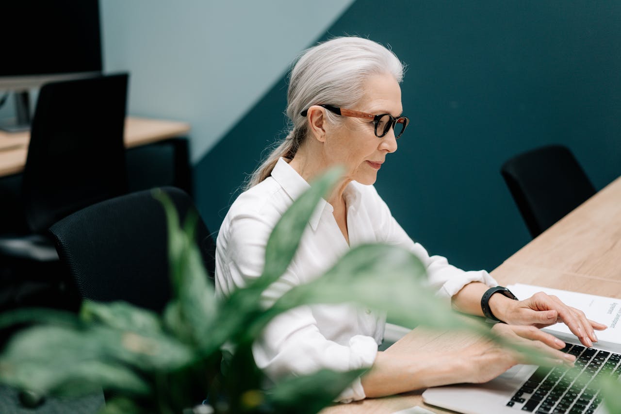 our-story Confident senior woman with glasses working on a laptop in a modern office setting.
